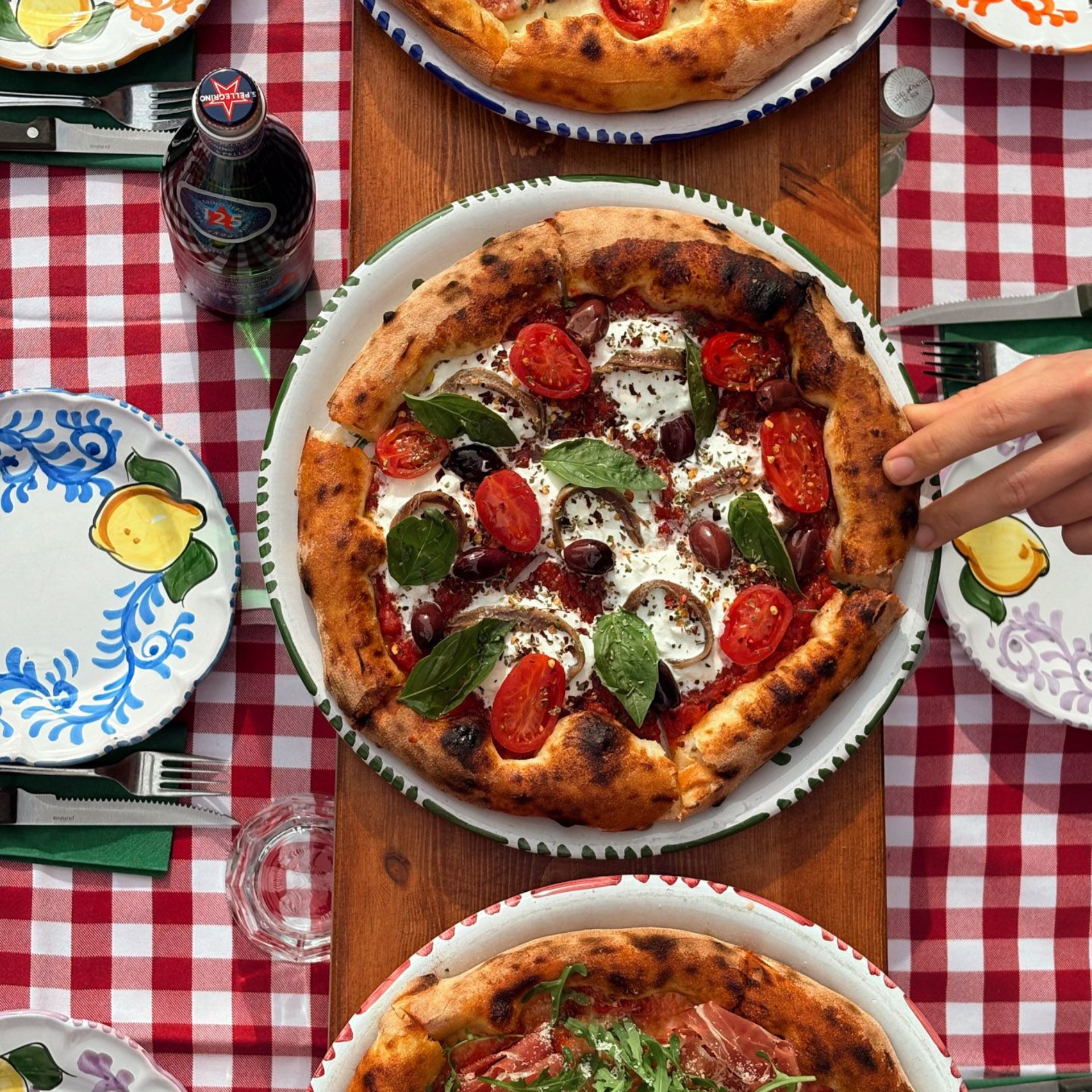 Pizza spread on a red checkered tablecloth with various plates, and a bottle of soda.
