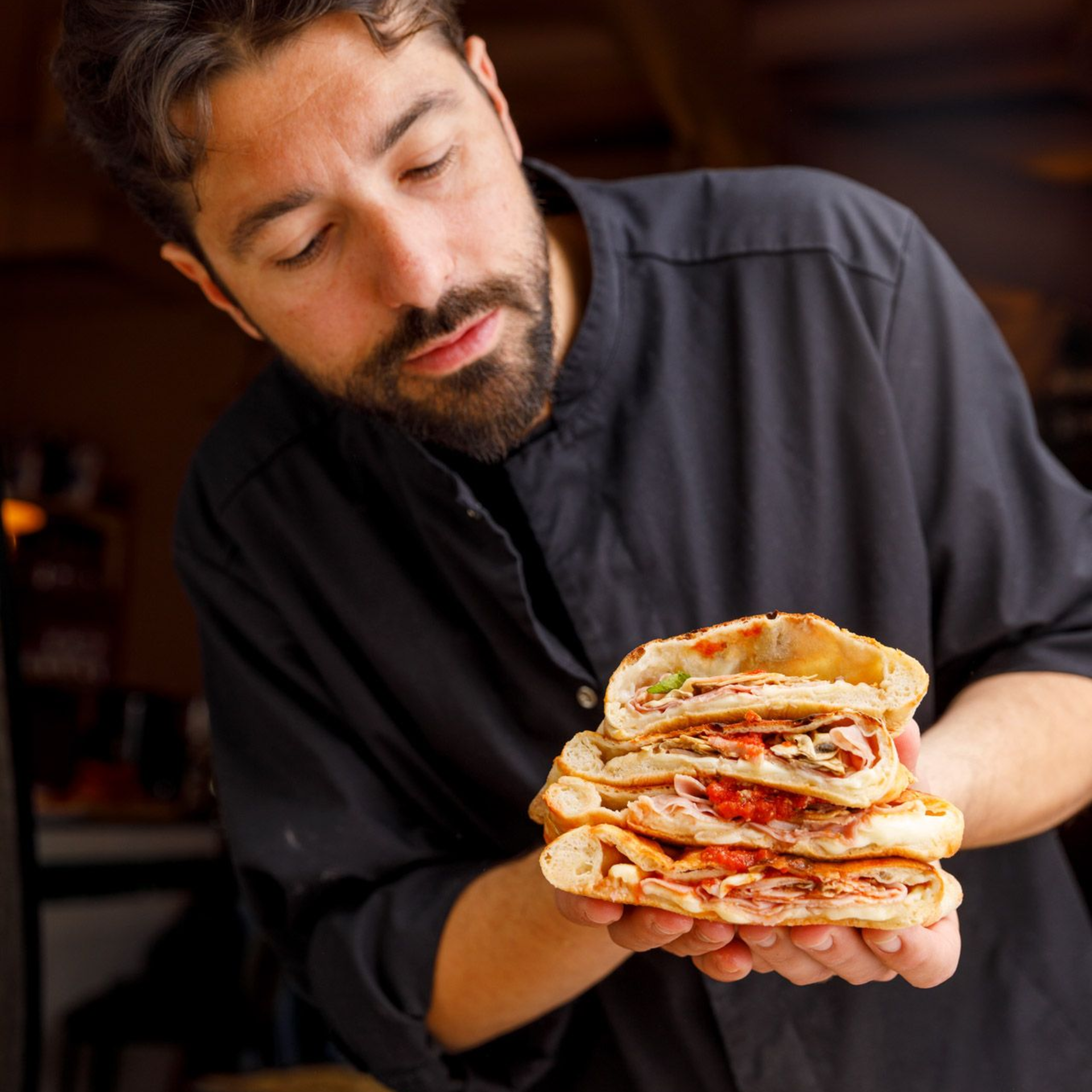Chef Luca holding a stack of layered sandwiches, inspecting them intently. Dark clothing, warm setting.