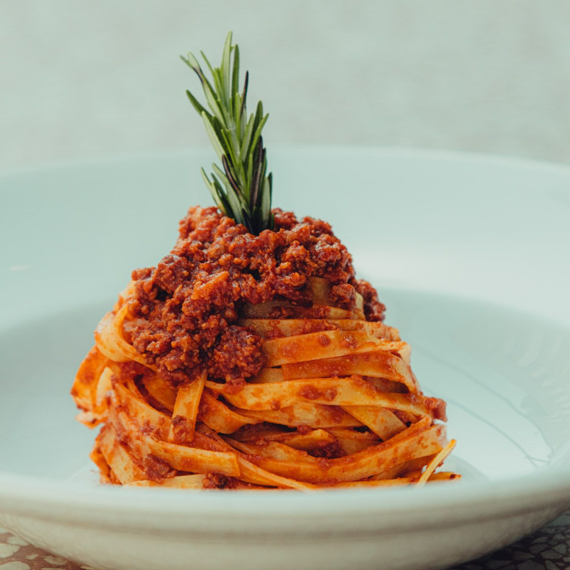 Fettuccine pasta with meat sauce, topped with rosemary sprig, served in a white bowl.