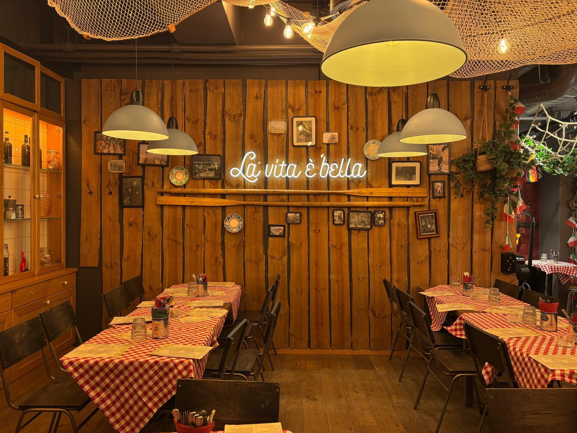 Restaurant interior with wooden walls, checkered tablecloths, and hanging lights; a neon sign reads 