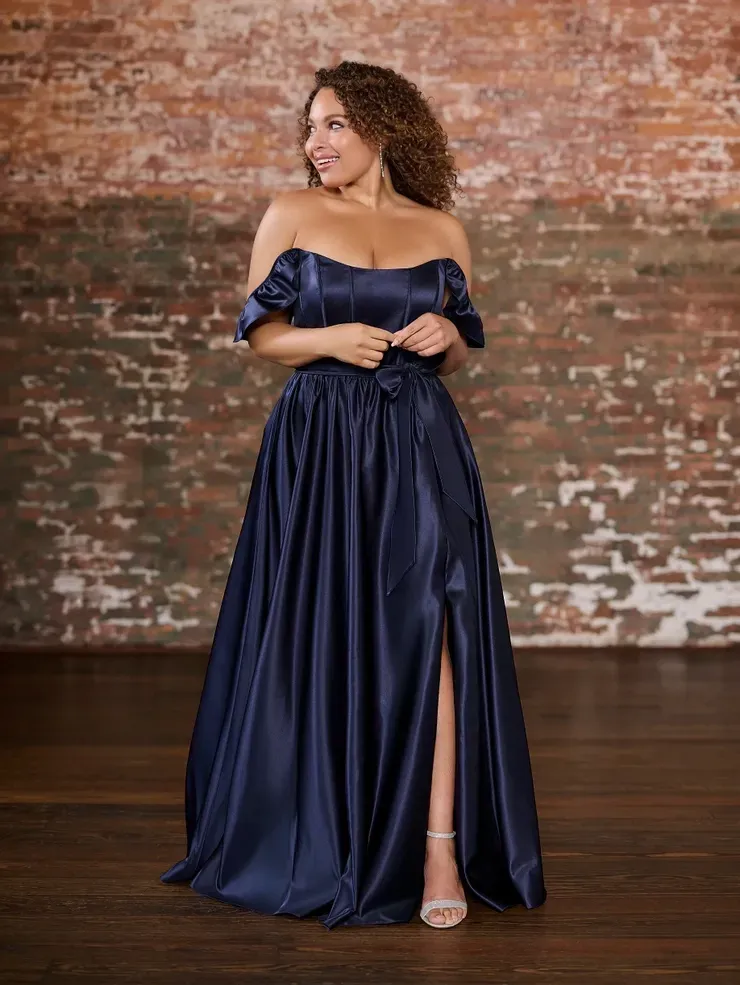 A woman in a blue satin off the shoulder dress is standing in front of a brick wall.