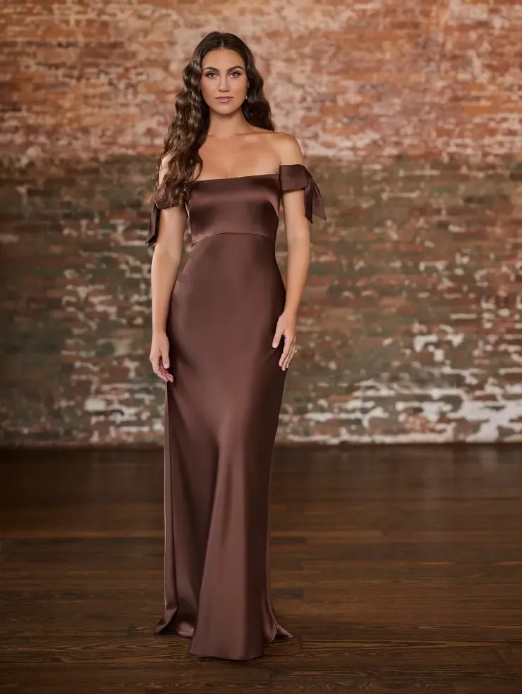 A woman in a long brown dress is standing in front of a brick wall.