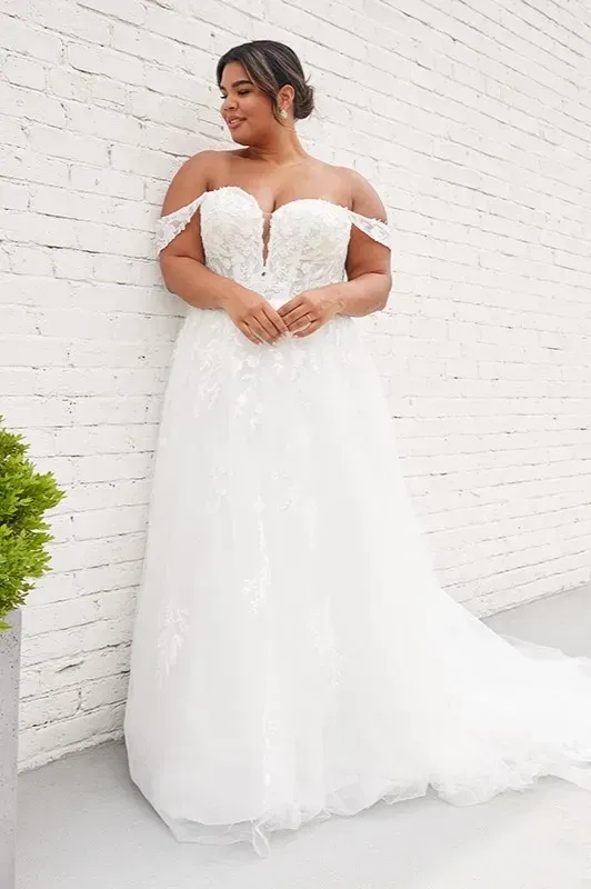 A plus size bride in a white wedding dress is standing in front of a white brick wall.