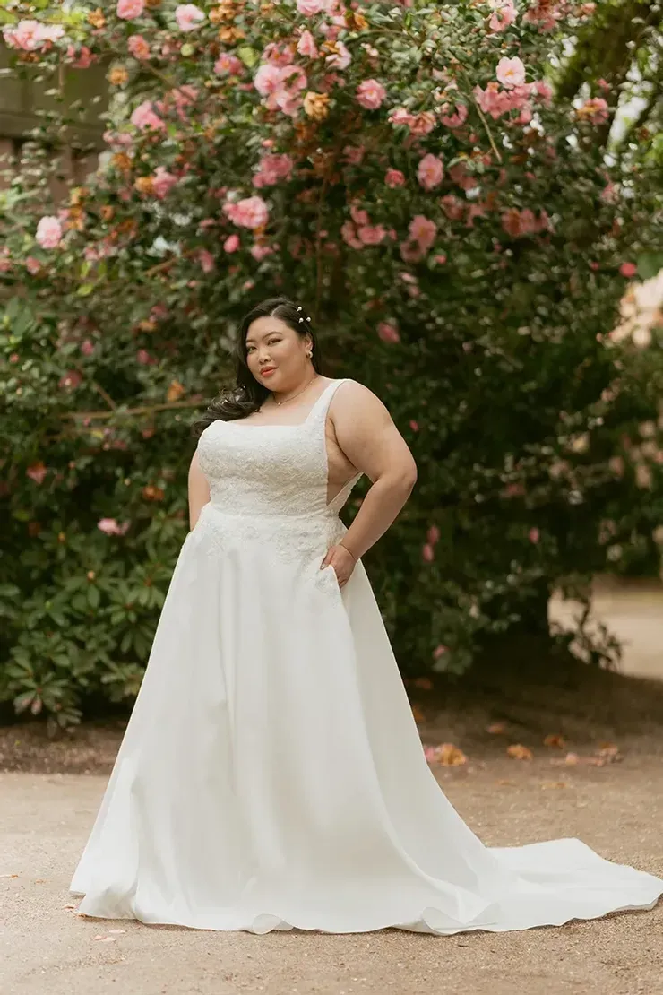 A plus size bride in a white wedding dress is standing in front of a tree.
