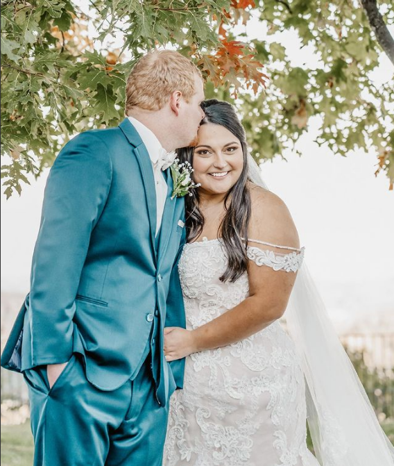 A bride and groom are kissing under a tree on their wedding day.