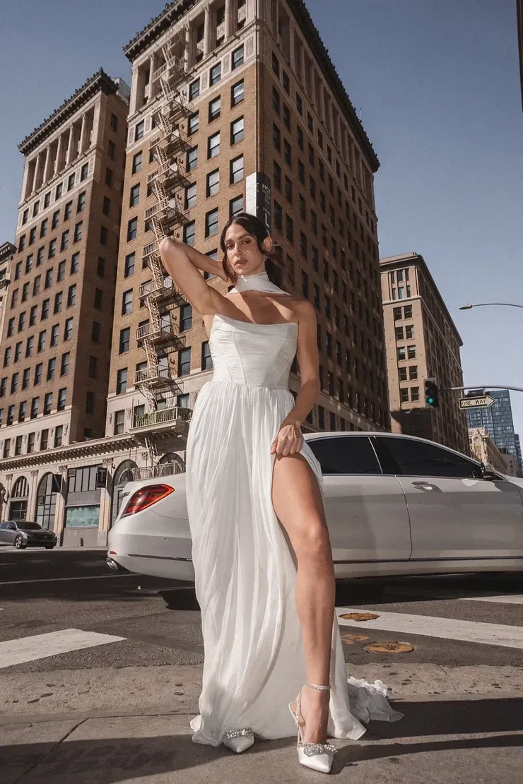 A woman in a white dress is standing in front of a building and a car.