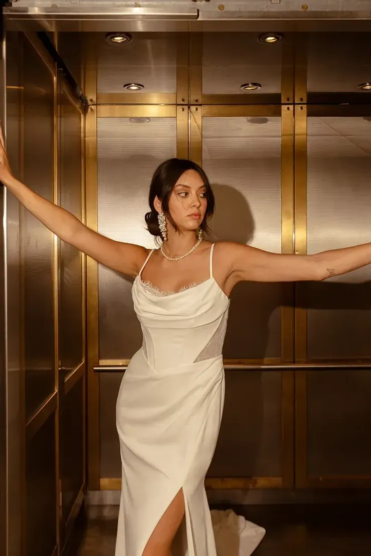 A woman in a white dress is standing in an elevator with her arms outstretched.