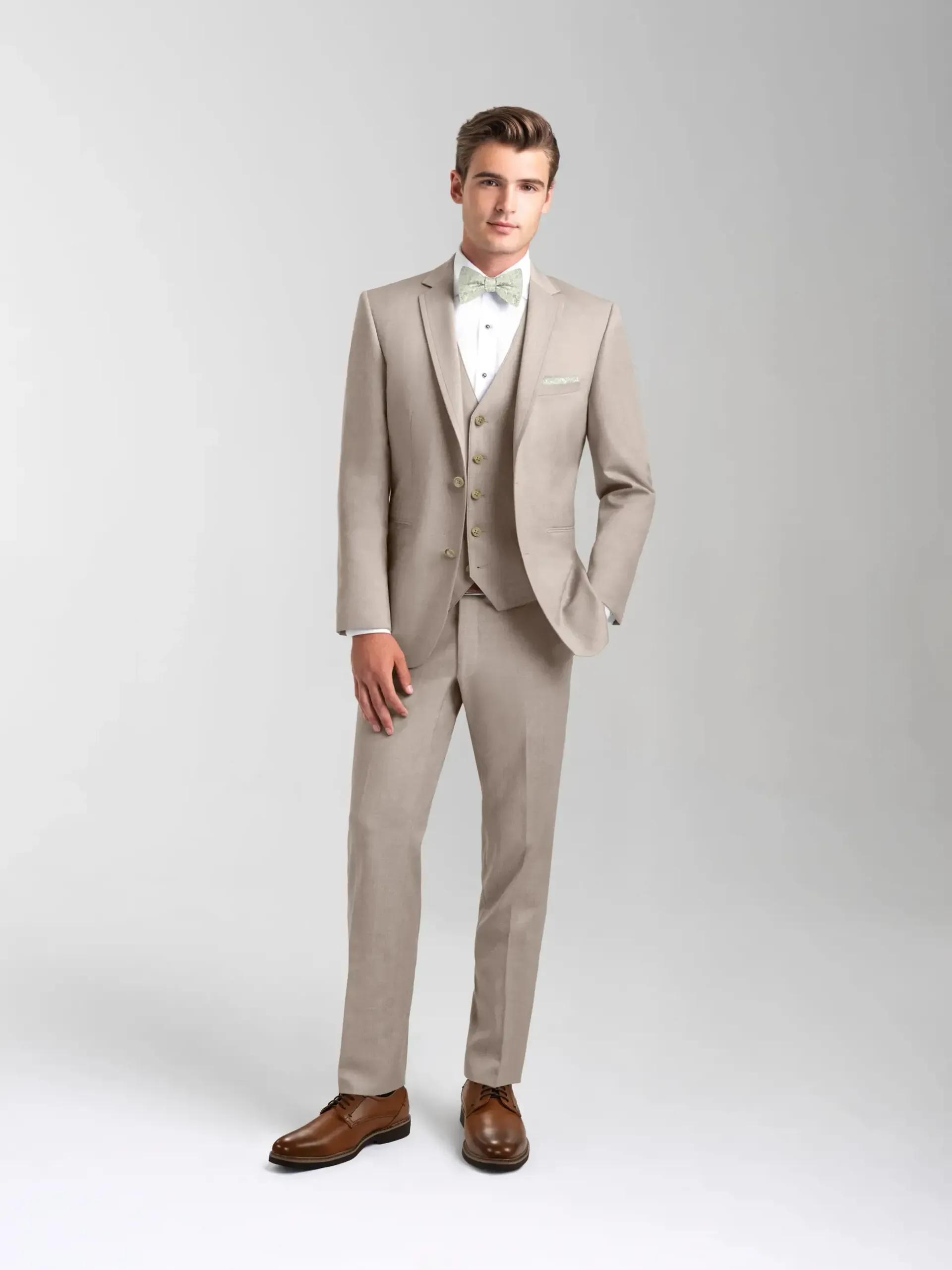A man in a tan suit and bow tie is standing on a white background.
