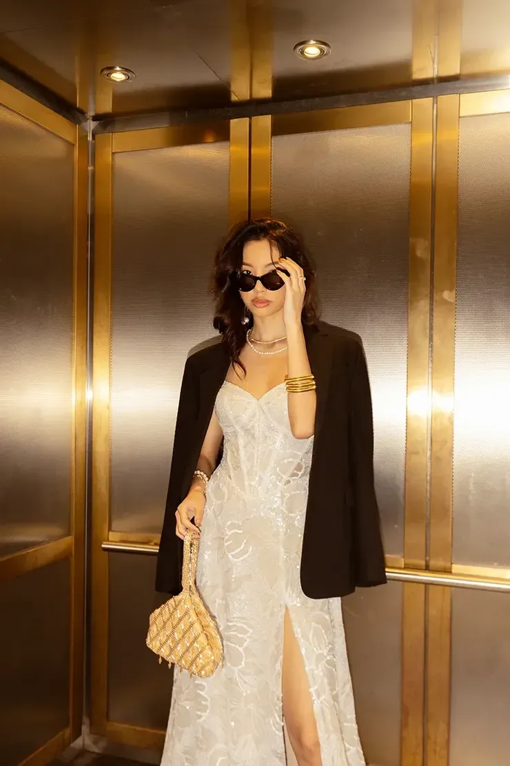 A woman in a white dress and black jacket is standing in an elevator.