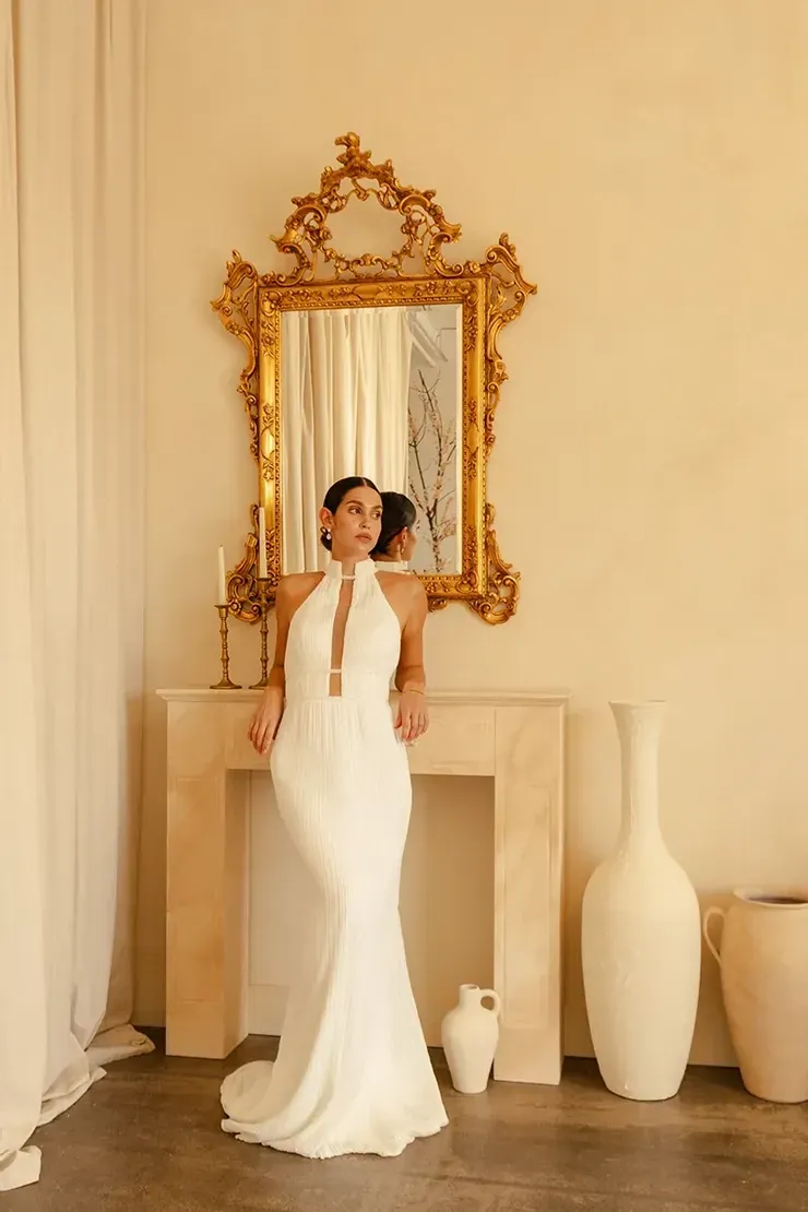 A woman in a white dress is standing in front of a mirror in a room.