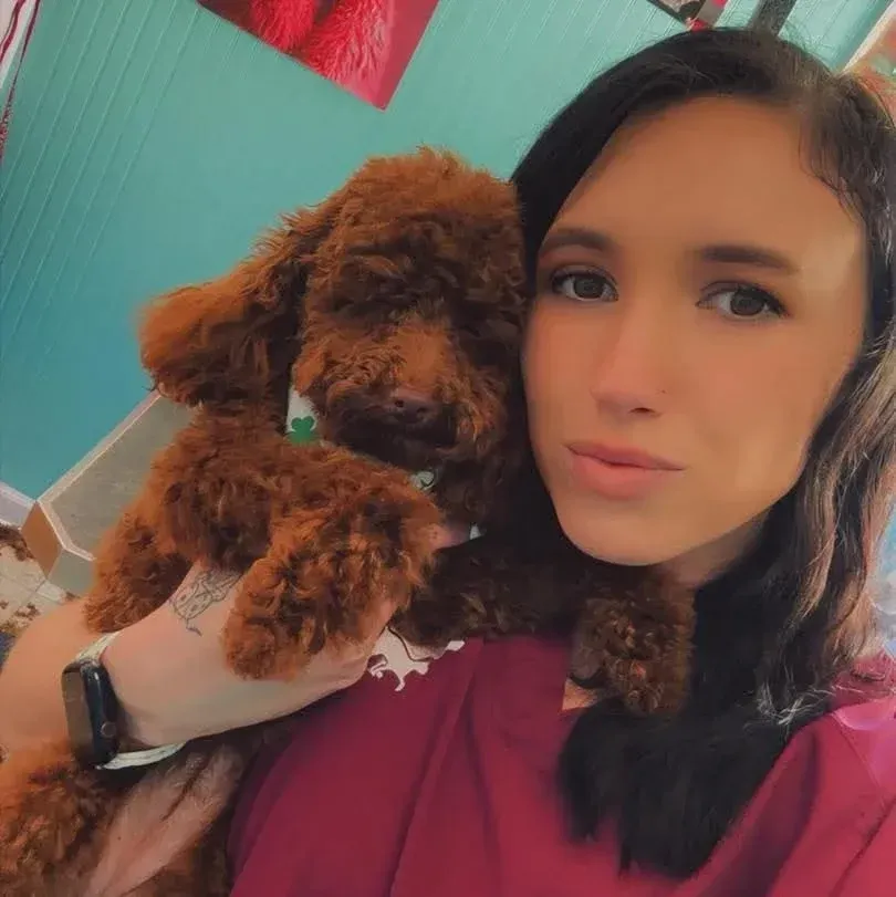 Woman holding a small curly brown dog, both close to the camera indoors.