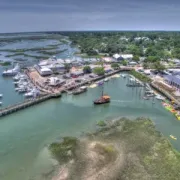 Aerial view of a coastal marina with boats, docks, waterfront buildings, and green wetlands nearby