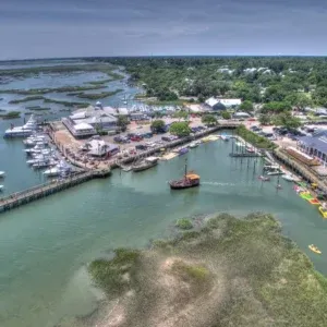 Aerial view of a coastal marina with boats, docks, waterfront buildings, and green wetlands nearby