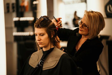 Une coiffeuse coupe les cheveux d'une cliente dans un salon, celle-ci étant assise sous une cape noire.