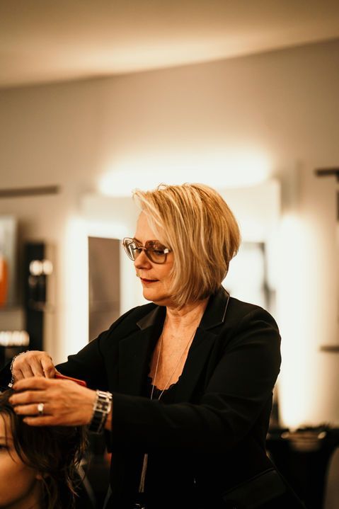 Une coiffeuse à lunettes travaille sur les cheveux d'une cliente dans un salon à l'éclairage tamisé.