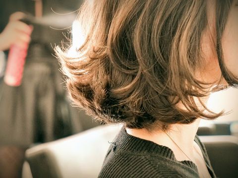 Vue de profil d'une coupe au carré courte et ondulée brune, dans un fauteuil de salon, avec un coiffeur tenant un fer à friser derrière elle.