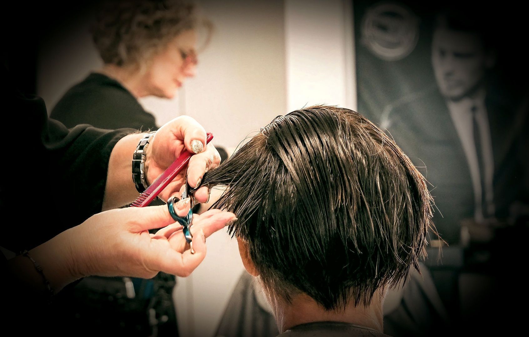 Une coiffeuse coupe les cheveux courts et foncés d'une cliente dans un salon de coiffure ; on aperçoit les ciseaux et le peigne.
