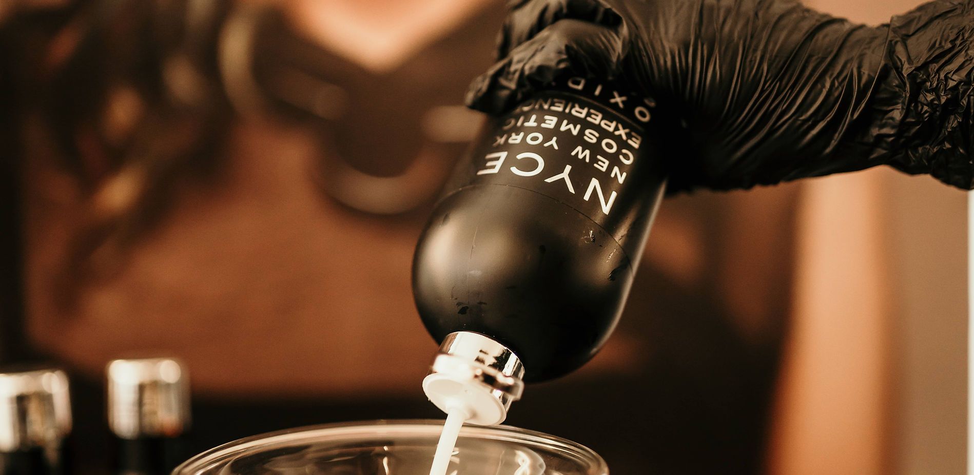 Main gantée versant un liquide transparent d'une bouteille noire dans un verre, sur fond brun chaud.