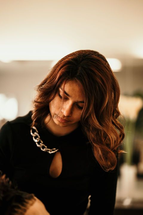 Femme aux cheveux bruns ondulés, regardant vers le bas, vêtue d'un haut noir et d'une chaîne en argent, dans une lumière intérieure chaleureuse.