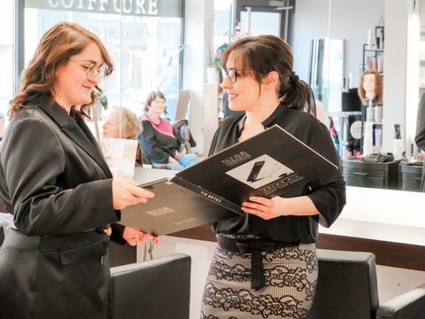Dans un salon de coiffure, deux femmes discutent d'un portfolio ; l'une d'elles tient de grands dossiers noirs et sourit.