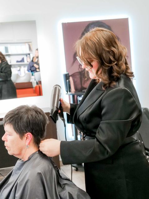 Une coiffeuse sèche les cheveux courts d'une cliente dans un salon, avec un miroir et des produits en arrière-plan.