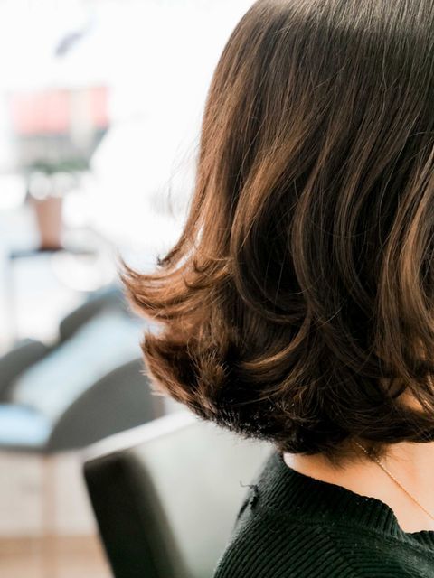 Gros plan sur des cheveux bruns mi-longs avec des boucles souples aux pointes, à l'intérieur, près d'un fauteuil de salon flou.