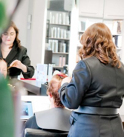 Une coiffeuse travaille sur les cheveux d'une cliente dans un salon lumineux, avec un miroir et des produits à portée de vue.