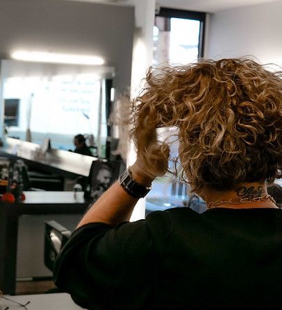 Personne aux cheveux bouclés ajustant sa coiffure devant un miroir de salon, vue de dos.