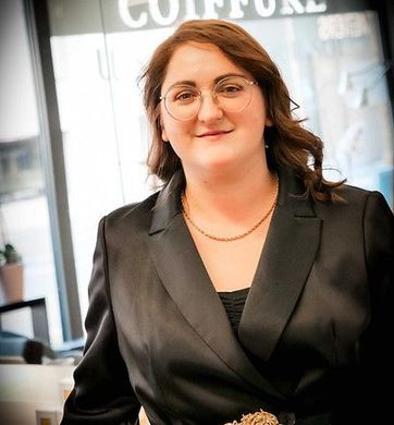 Femme en blazer noir souriant à l'intérieur, debout devant la vitrine d'un magasin.