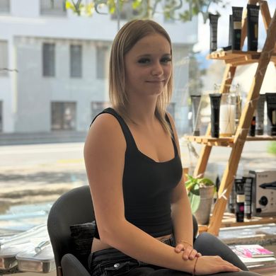 Une femme assise près d'une fenêtre, vêtue d'un débardeur noir, avec des étagères et une rue de la ville visibles derrière elle.