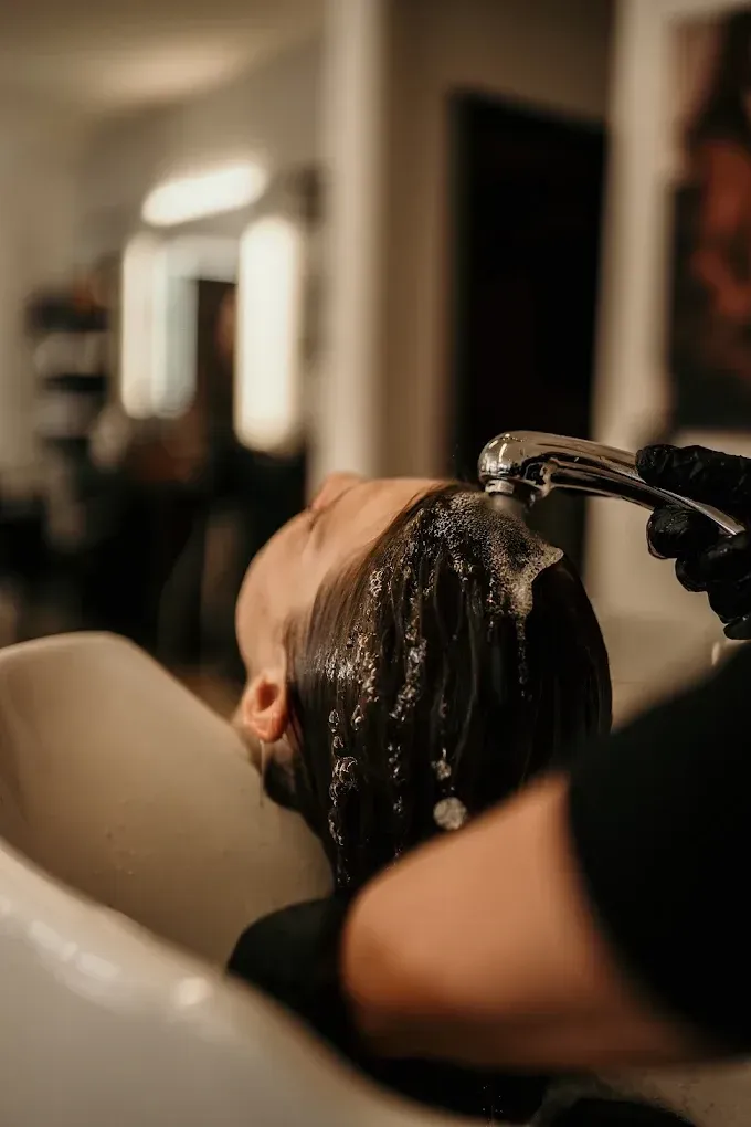 Des cheveux sont rincés au lavabo d'un salon de coiffure par un coiffeur sous une lumière chaude.