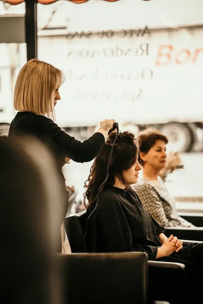 Une coiffeuse boucle les cheveux d'une cliente assise dans un salon lors d'une séance de coiffure.