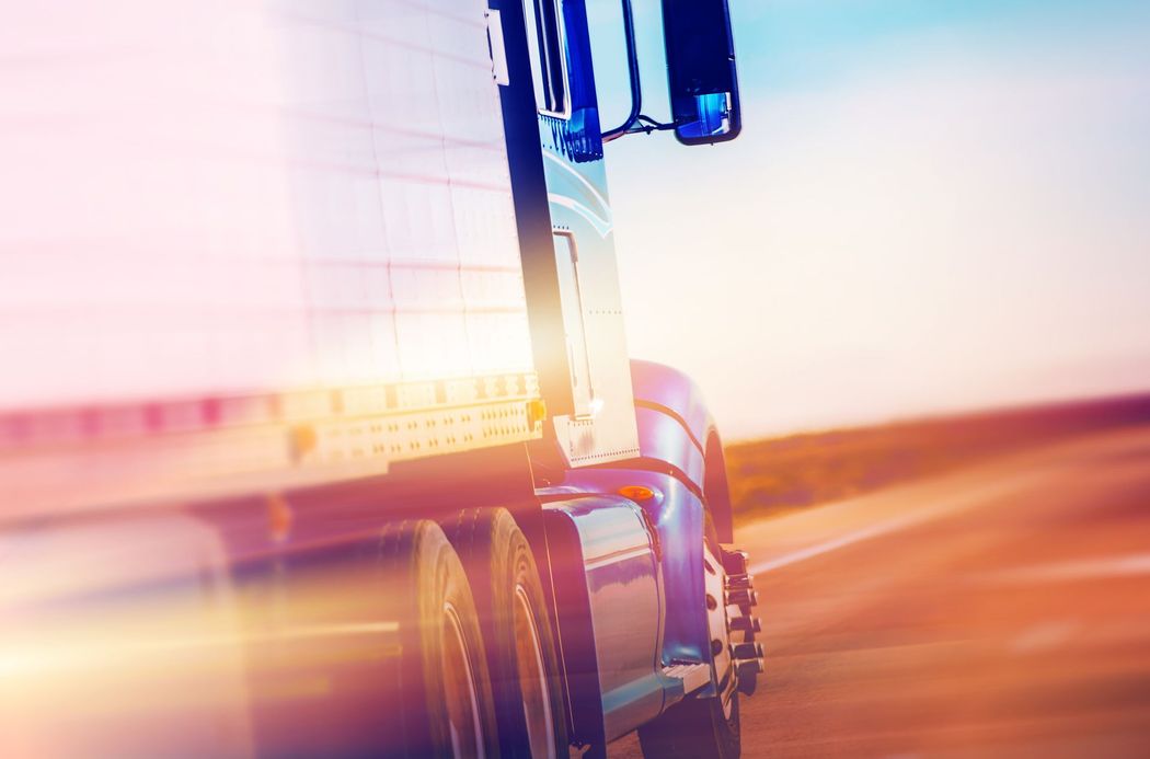 Camion blu che percorre una strada al tramonto con effetto mosso e calda luce arancione.