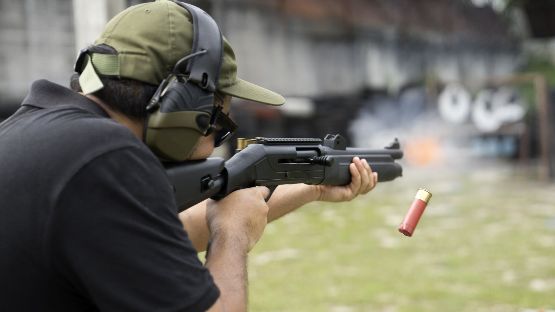 Man firing a shotgun at a target, ejection of a shell visible. Wearing hearing protection, outdoors.