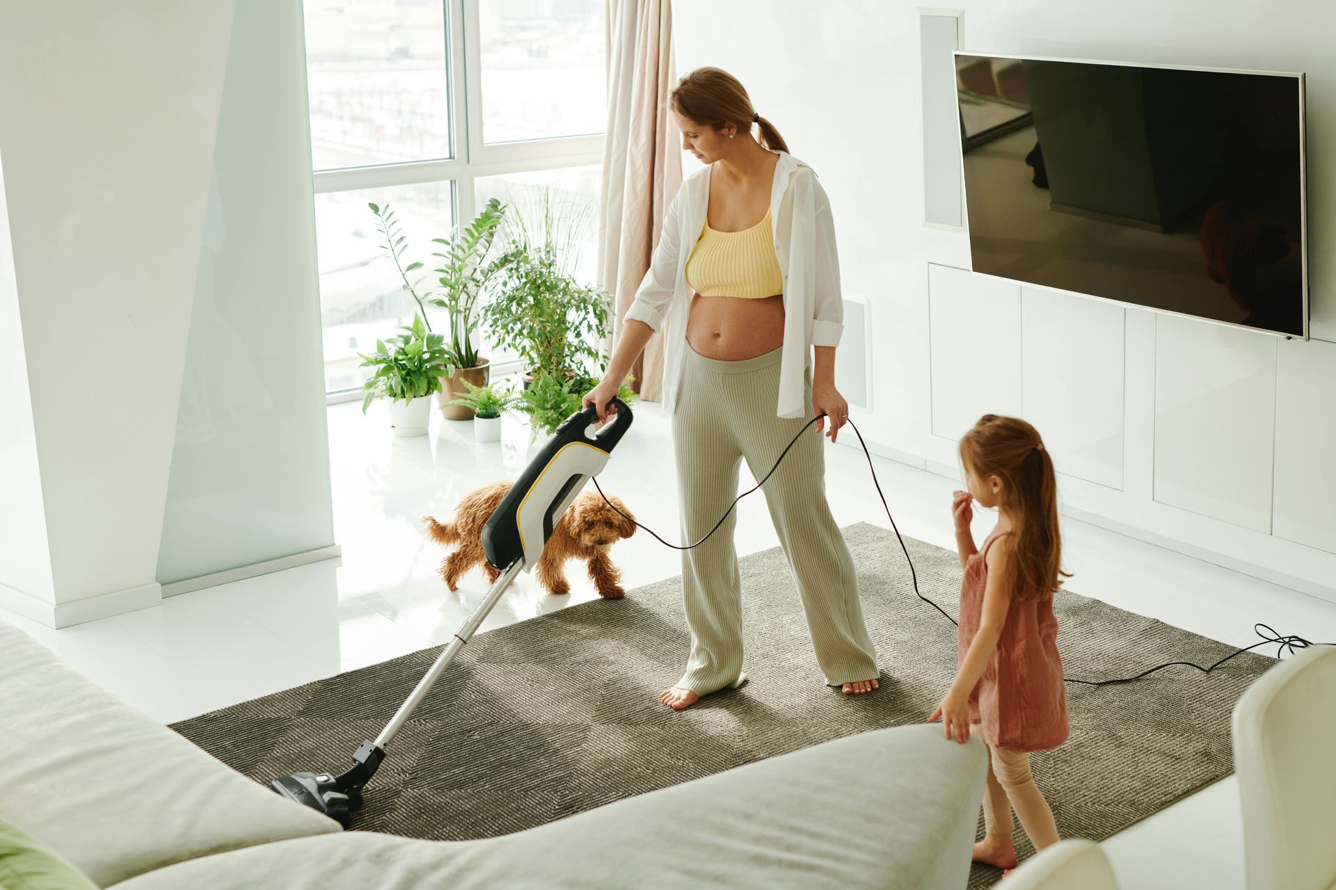 A pregnant woman is cleaning the floor with a vacuum cleaner while a little girl walks her dog.