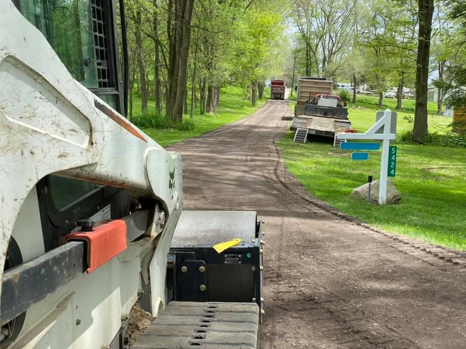 Tree-lined path beside construction vehicle, green sign, and small maintenance trailer near a park road