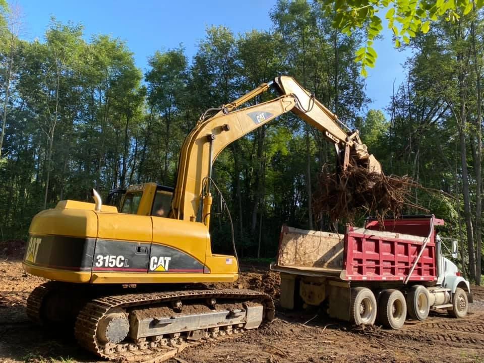 Yellow excavator loading soil into a red dump truck on a wooded site.
