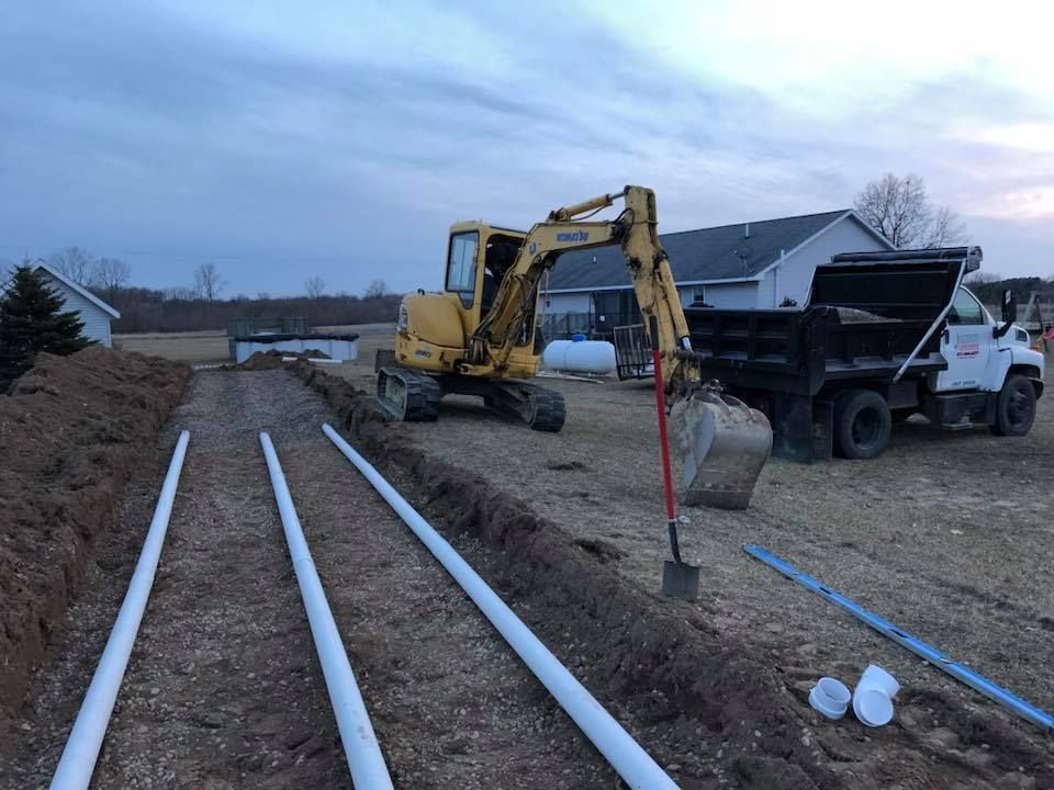 Excavator and dump truck beside a trench with white pipes laid along a dirt road at dusk