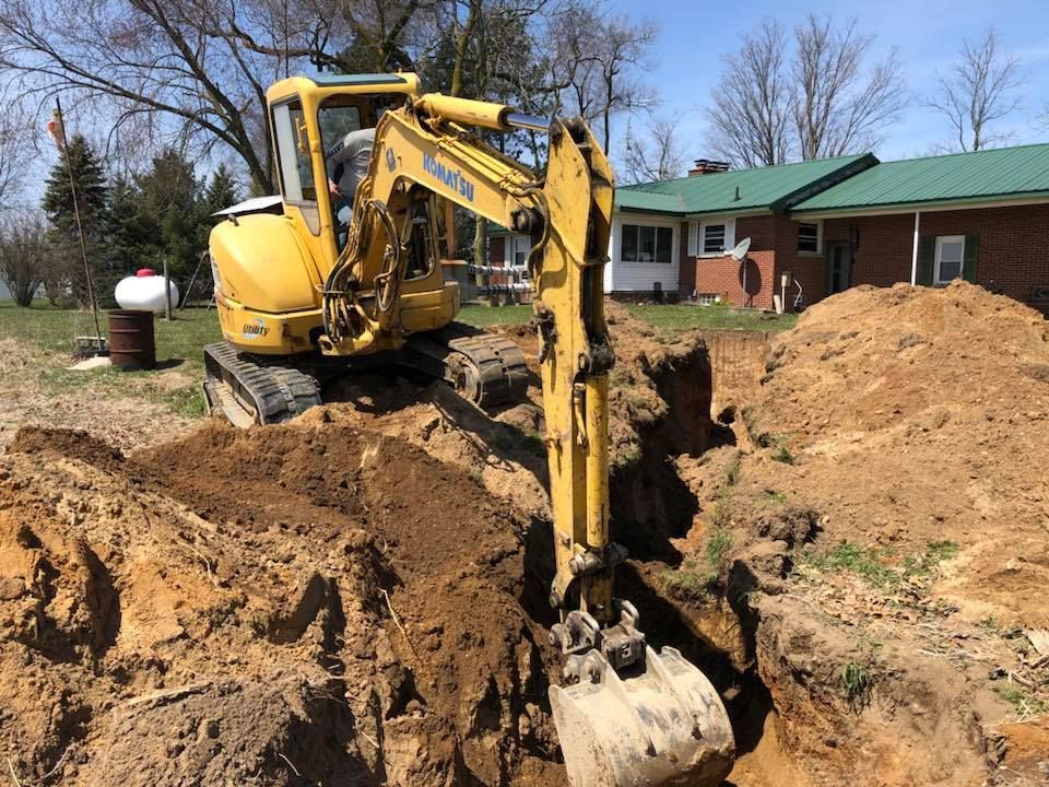 Yellow excavator digging a trench beside a brick house with dirt piles in a yard