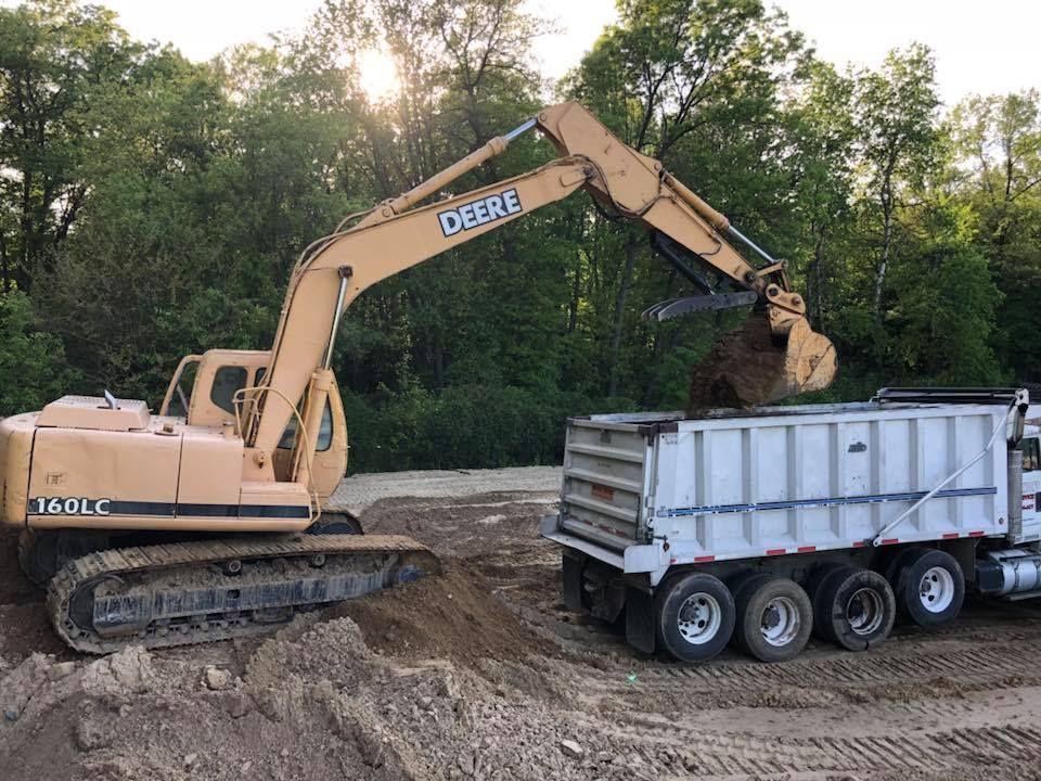 Excavator loading dirt into a white dump trailer at a wooded construction site