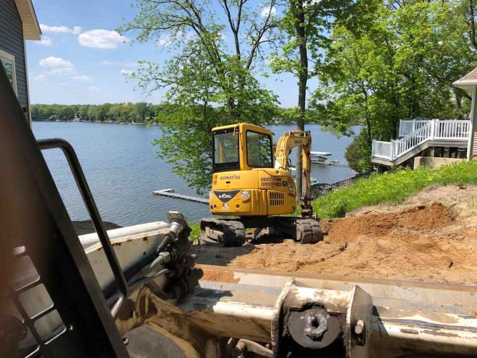 Yellow excavator on a lakeshore beside a house, digging dirt near the water on a sunny day