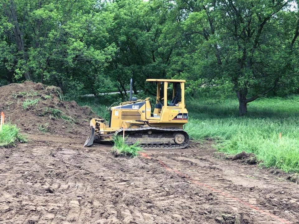 Yellow bulldozer clearing dirt in a wooded area with trees and grass nearby
