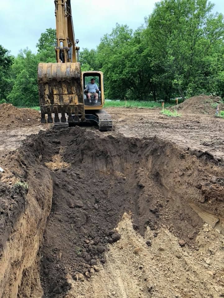 Excavator digging a large pit in a dirt clearing with green trees in the background.