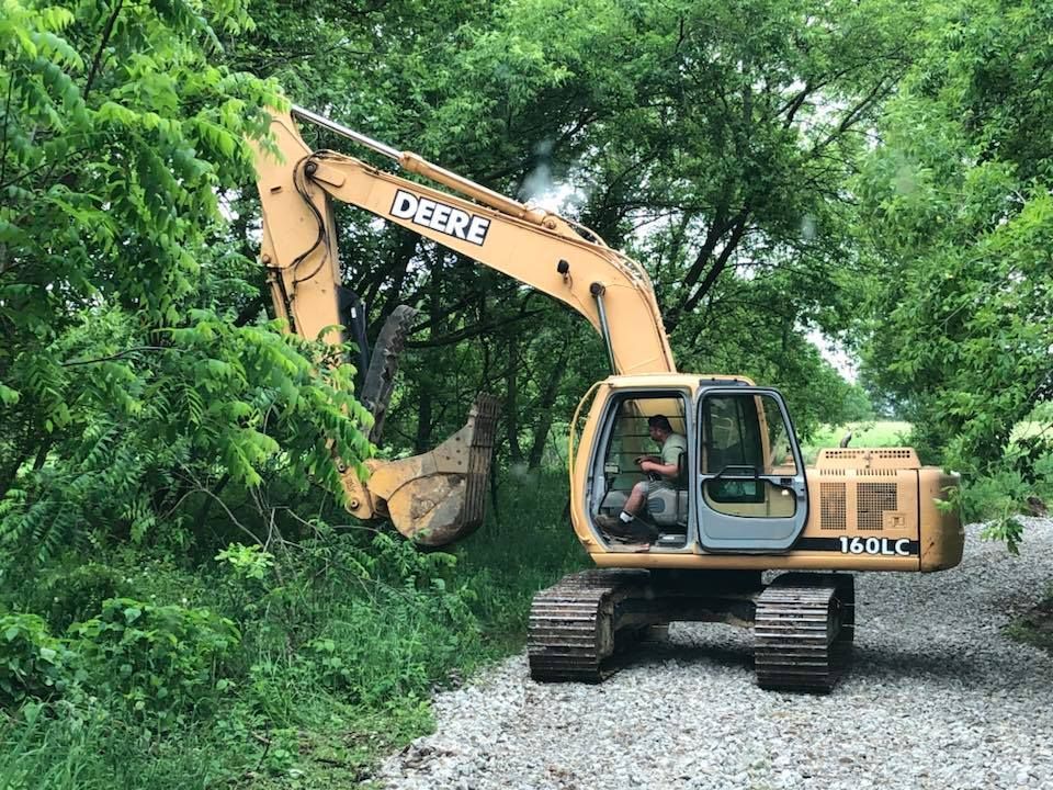 Yellow John Deere excavator clearing brush beside a gravel road under green trees