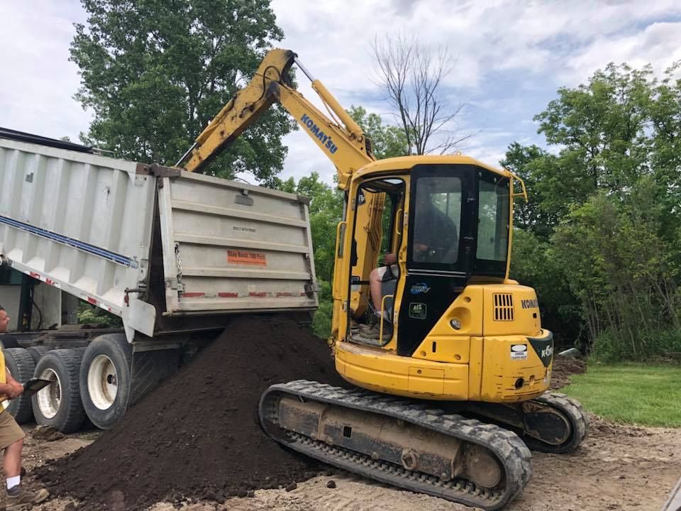 Yellow excavator unloading dark soil from a dump trailer outdoors.