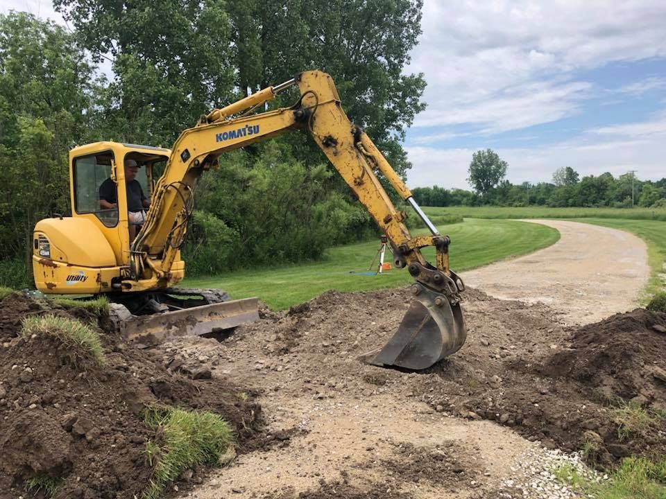 Yellow excavator digging along a dirt path beside grassy field and trees