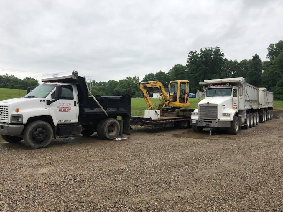 Two dump trucks and a yellow excavator parked on a gravel lot under cloudy skies