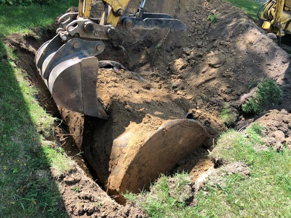 Excavator bucket lifting a large rock from a trench beside a grassy area