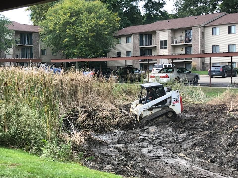 Small white skid-steer excavator clearing muddy ground beside an apartment building and tall grass.
