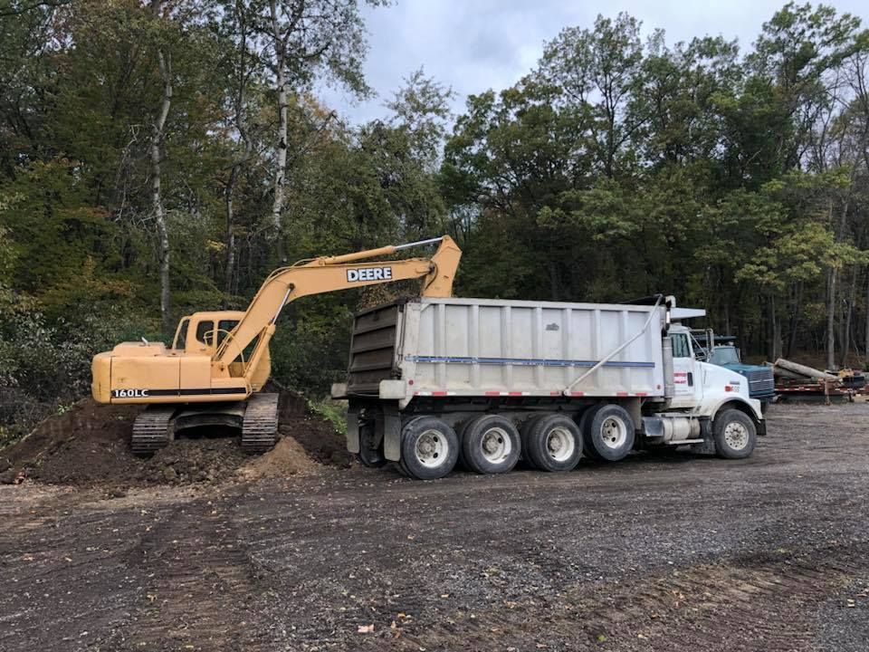 Yellow excavator loading soil into a white dump truck at a wooded worksite